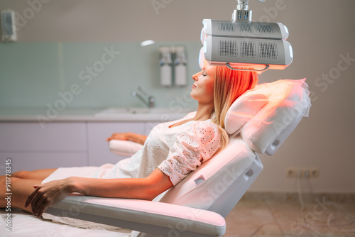 Relaxed blonde female with closed eyes lying on procedure chair having LED red light hair photodynamic therapy during hair treatment at clinic. Woman client having non-invasive type of phototherapy.