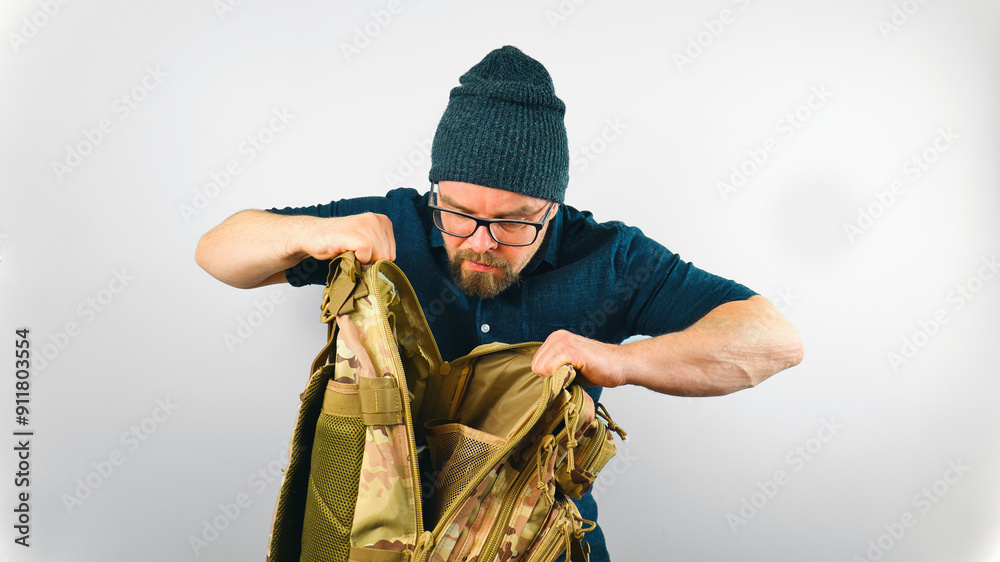 Portrait of handsome bearded man with beanie and glasses looking into camo military backpack, isolated on white background