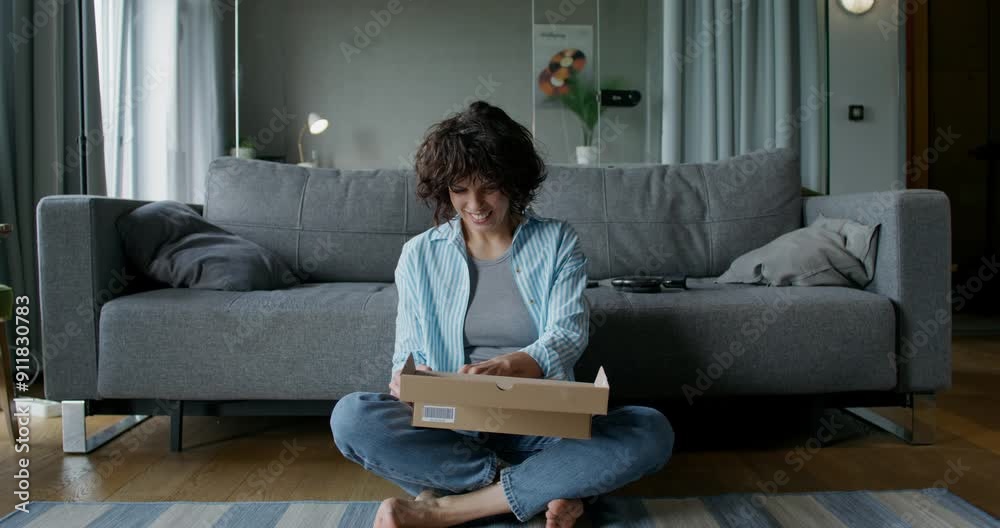 The woman opens the box and pulls out the cosmetics, she smiles. She is sitting on the floor near the sofa at home. Daylight The woman opens the box and pulls out the cosmetics, she smiles. She is sitting on the floor near the sofa at home. Daylight