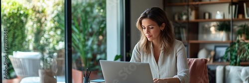 A woman sitting at her desk in front of an open laptop, working on marketing for a small business. In the background is a modern interior design office with a glass door leading to an outdoor area.