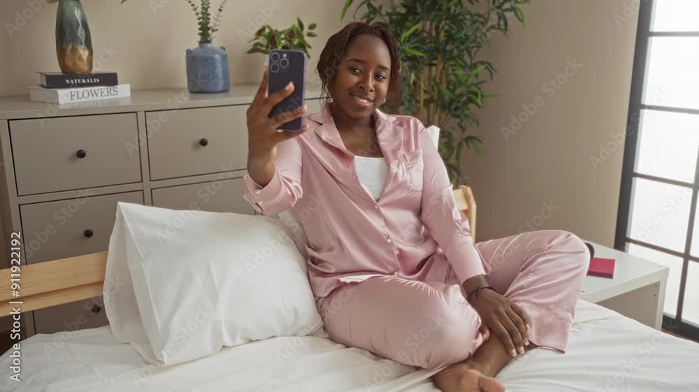 Young african american woman sitting on a bed in her bedroom taking a selfie with her smartphone in a relaxed home environment Young african american woman sitting on a bed in her bedroom taking a selfie with her smartphone in a relaxed home environment