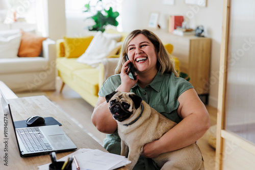 Happy woman hugging pug dog while talking on phone at home office photo