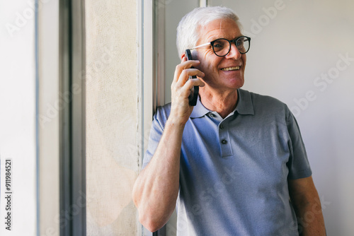 Happy senior man in glasses talking on cellphone near window photo