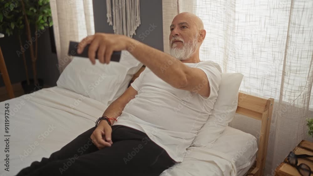 Mature bald man with grey beard relaxing in bed at home with a remote control in hand in a cozy bedroom Mature bald man with grey beard relaxing in bed at home with a remote control in hand in a cozy bedroom