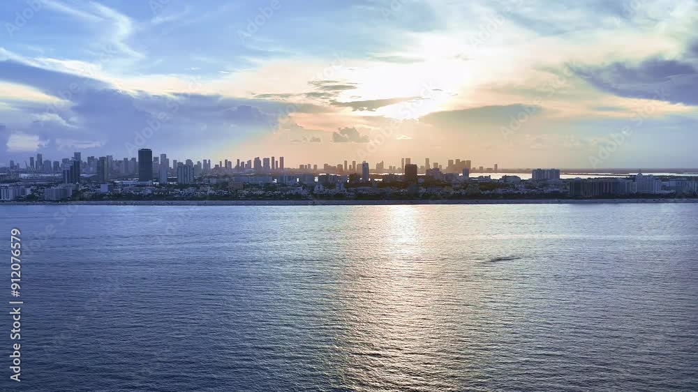 A drone flies toward Miami Beach from the Atlantic Ocean at sunset, capturing the sea, skyline, and shoreline, ending near the beach. A drone flies toward Miami Beach from the Atlantic Ocean at sunset, capturing the sea, skyline, and shoreline, ending near the beach.