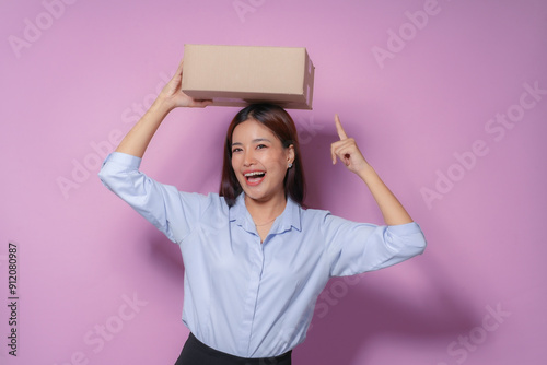 Young woman holding cardboard box on head, pointing up with index finger and laughing, isolated on purple background