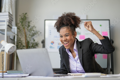 Young office worker is raising her fist in the air, excited about a win she just had while working on her laptop