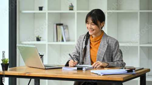 An attractive young businesswoman doing some paperwork and using laptop in a modern office