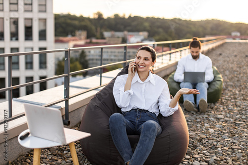 Two colleagues working remotely on rooftop, using laptops and phones. Casual work environment promotes relaxation and productivity. Outdoors setting emphasizes flexibility and modern corporate culture