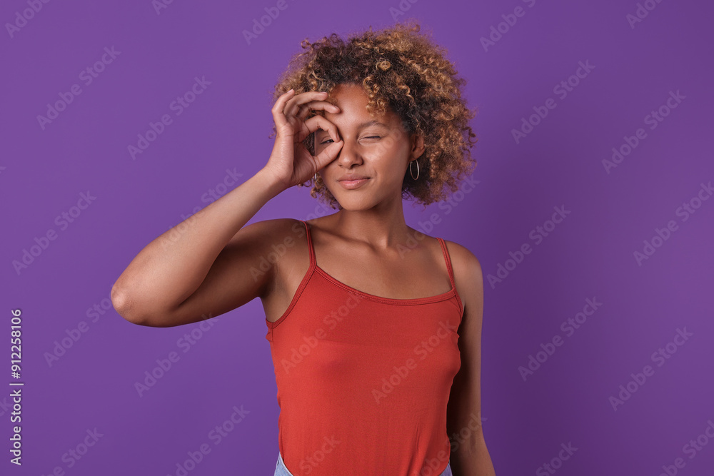 A young woman with curly hair playfully poses against a vibrant purple backdrop, showcasing a fun expression while winking and forming a playful gesture with her hand. A young woman with curly hair playfully poses against a vibrant purple backdrop, showcasing a fun expression while winking and forming a playful gesture with her hand.