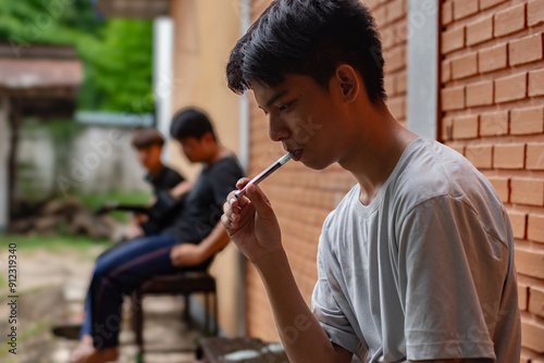 Asian schoolboy sitting near brick wall and smoking e-cigarette while his frineds looking at him doubtfully, teenagers' problem and curiosity to try concepts. photo