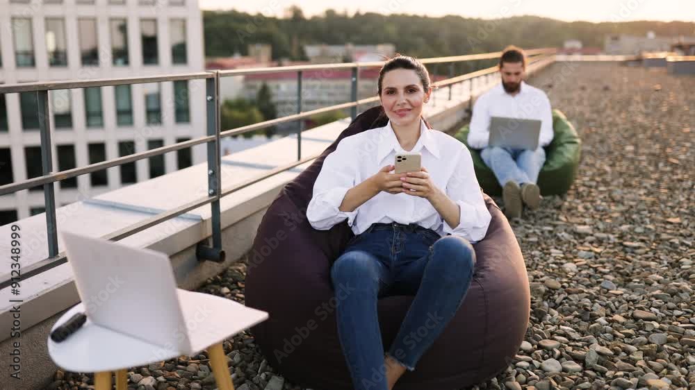 Young woman sitting on rooftop terrace using smartphone and giving thumbs up. Casual and relaxed atmosphere, enjoying outdoor setting in comfortable bean bag chair. Happy and content expression. Young woman sitting on rooftop terrace using smartphone and giving thumbs up. Casual and relaxed atmosphere, enjoying outdoor setting in comfortable bean bag chair. Happy and content expression.