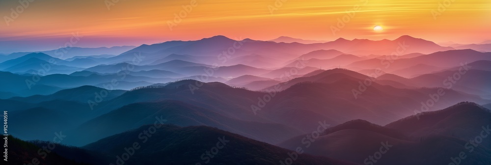 Fototapeta premium The mountains are covered in a misty blue haze. The sun is setting in the distance, casting a warm glow over the landscape. The sky is a mix of orange and pink hues, creating a serene