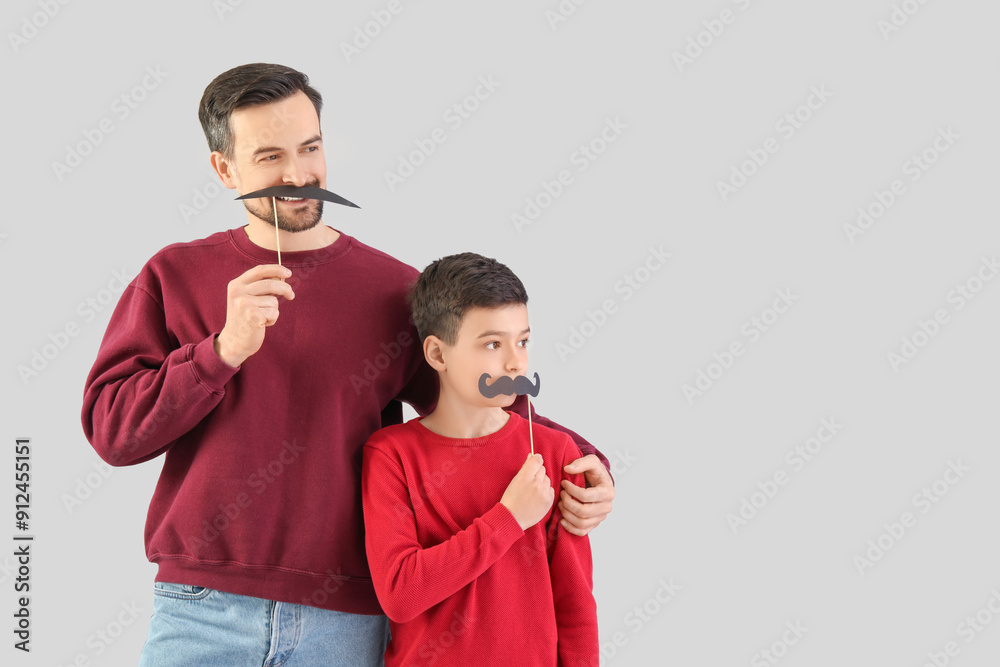 Portrait of father and his little son with paper mustache on light background Portrait of father and his little son with paper mustache on light background