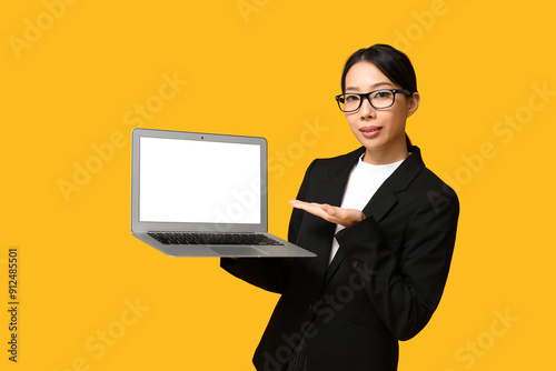 Portrait of young Asian businesswoman showing modern laptop with blank screen on yellow background