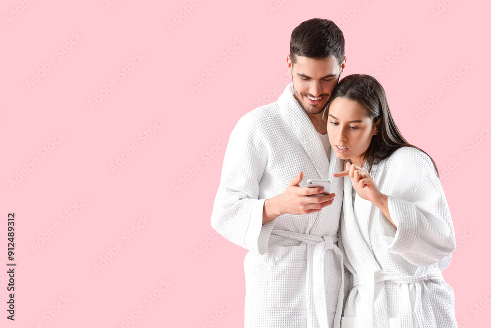 Happy young couple in robes with mobile phone on pink background Happy young couple in robes with mobile phone on pink background