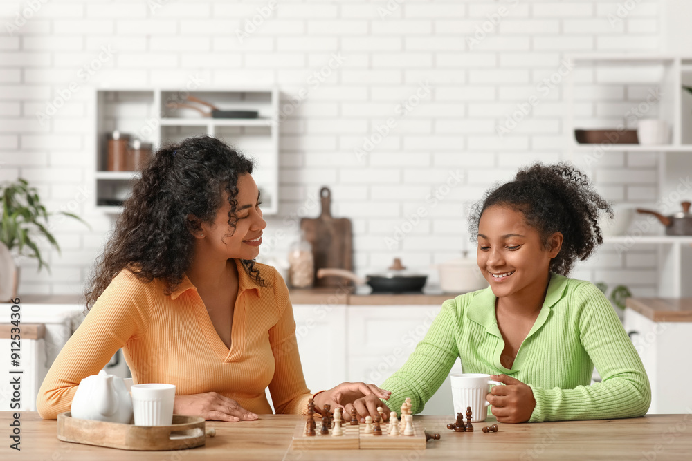 Beautiful young happy African-American mother with her daughter playing chess in kitchen at home Beautiful young happy African-American mother with her daughter playing chess in kitchen at home
