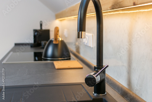 Detail of black sink faucet in modern kitchen with black countertop, white Venetian plaster wall and wooden led backlighting 