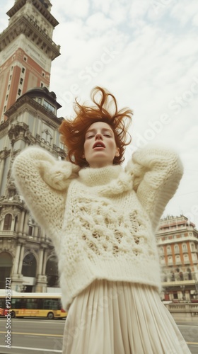 A woman in a thick, knit sweater stands with a cityscape backdrop, striking a relaxed pose in an urban environment. photo