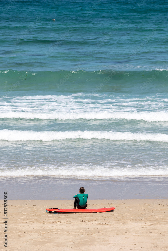 A person in a wetsuit sits on the beach, leaning against a red surfboard and gazing out towards the rolling waves of the ocean, embodying peace, reflection, and solitude. A person in a wetsuit sits on the beach, leaning against a red surfboard and gazing out towards the rolling waves of the ocean, embodying peace, reflection, and solitude.