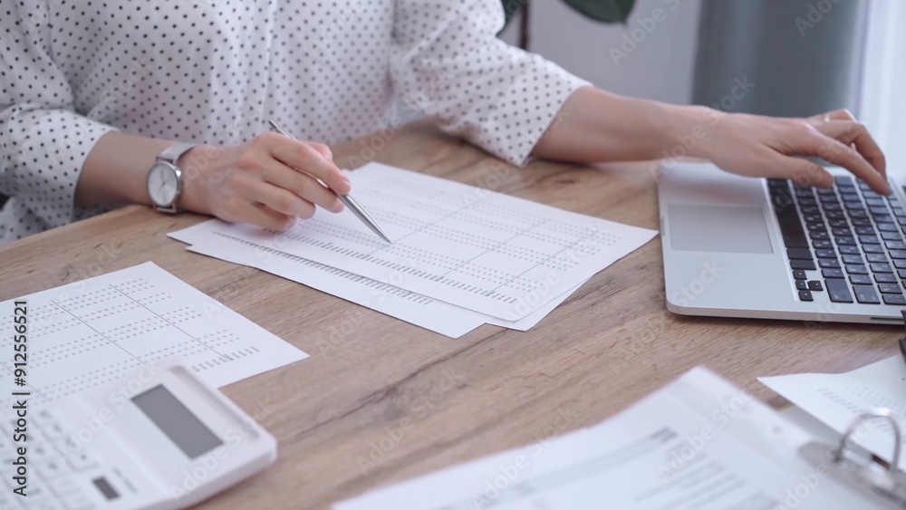 Fototapeta premium Close-up of a woman's hand checking financial paper beside a laptop on the wooden desk. Bookkeeping, finance audit and taxes