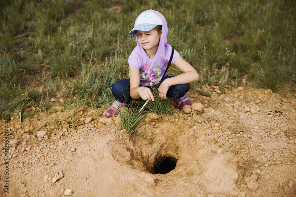 A girl in nature at a rodent burrow sits in a cap and hood. Young researcher A girl in nature at a rodent burrow sits in a cap and hood. Young researcher