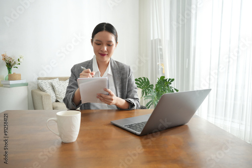 Картината върху платно An Asian woman takes notes while working from home, sitting at a wooden desk in a bright living room
