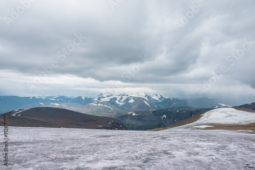 Dramatic panoramic view from big glacier to wide alpine valley and large snow-capped mountain range in rainy low clouds. Awesome vast landscape with high snowy mountains in rain under gray cloudy sky.