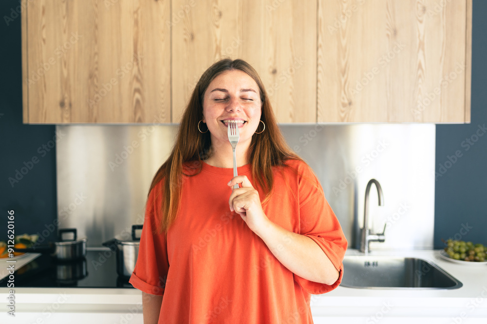 Portrait of excited hungry young woman holding fork in her mouth, biting and dreaming about delicious healthy food at the kitchen. Diet concept