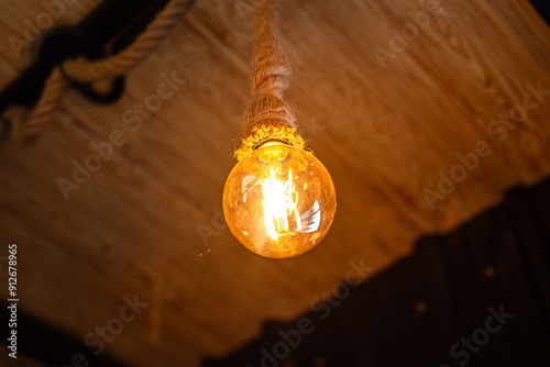 Rope ceiling lighting bulb glowing in orange warmlight shade, which is installed at living room for cozy design. Interior decoration object photo, close-up and selective focus. photo