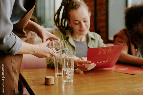 Fototapet Server pouring water into glass for guest reading menu in restaurant with wooden tables and cozy ambiance