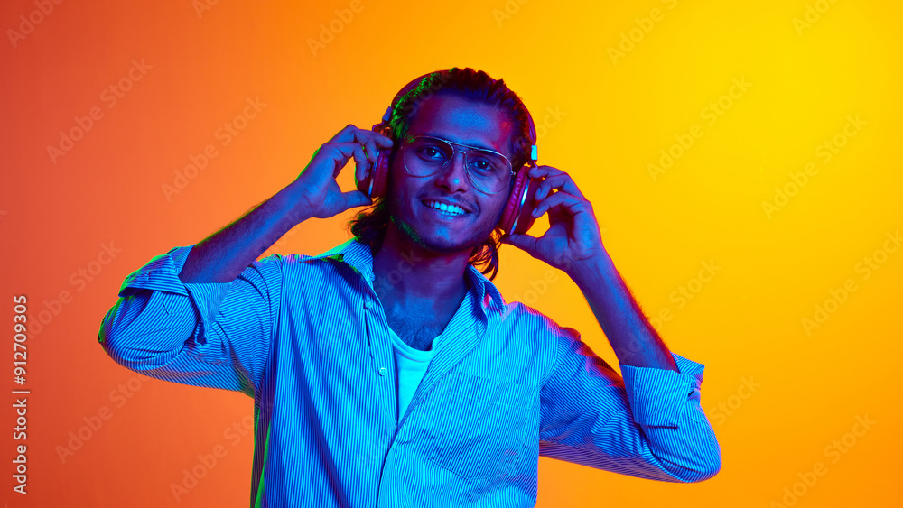 Young middle eastern man wearing headphones looking at camera to listen music in neon light against gradient orange background. Concept of human emotions, diversity, youth, modernity. Ad Young middle eastern man wearing headphones looking at camera to listen music in neon light against gradient orange background. Concept of human emotions, diversity, youth, modernity. Ad