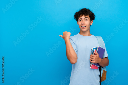 Quadro su tela Amazed boy guy high school male pupil student teenager with bag holding books copybooks pointing finger empty copy space for advertisement isolated on blue background