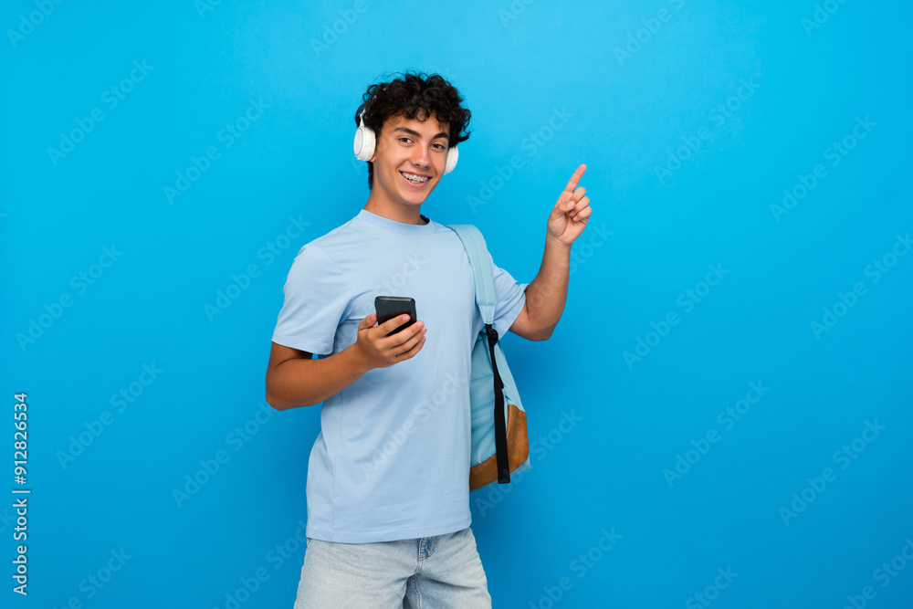 Smiling boy guy high school male pupil student teenager with backpack using smart phone listening music in headphones pointing finger up copy space isolated on blue background. Education concept Smiling boy guy high school male pupil student teenager with backpack using smart phone listening music in headphones pointing finger up copy space isolated on blue background. Education concept