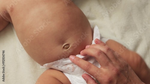 Unrecognizable mother massages newborn baby's tummy, gently touching child's navel, infant kid with naked body wearing only in diaper lying on white sheet
