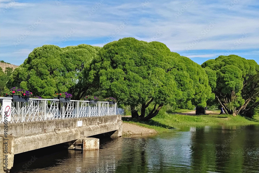 Charming bridge decorated with flowers spans over river, with lush green trees in background under partly cloudy sky Charming bridge decorated with flowers spans over river, with lush green trees in background under partly cloudy sky