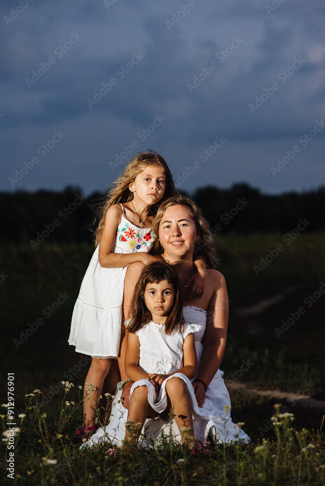 A mother and two daughters pose in white dresses at sunset. Family on vacation outside the city. Portrait of a woman with two children in nature. The concept of Mother's Day is family. A mother and two daughters pose in white dresses at sunset. Family on vacation outside the city. Portrait of a woman with two children in nature. The concept of Mother's Day is family.