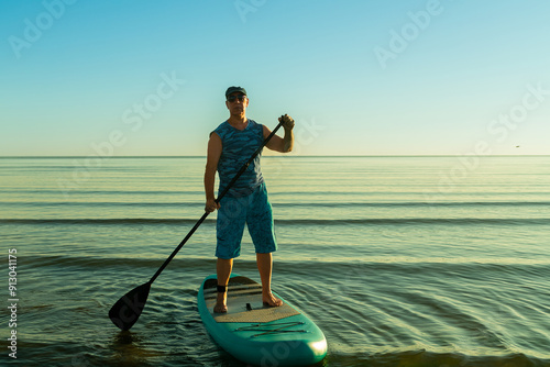 Man in shorts and sunglasses on a paddleboard with a paddle in the sea against the background of the dawn sky photo