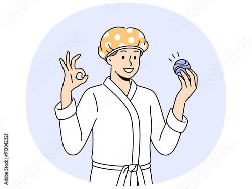 Man with bath bomb is dressed in bathrobe and bandage to protect hair from moisture. Young guy shows OK gesture recommending use of aromatic bath bomb while performing hygiene procedures.