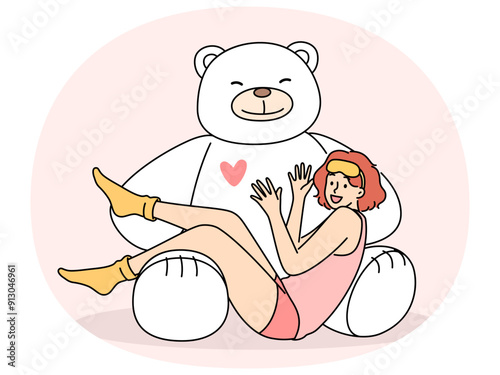 Giant teddy bear near sitting teenage girl in pajamas and sleep mask. Funny schoolgirl teen age looks at screen lying on feet of toy bear rejoicing at cute gift from parents or boyfriend