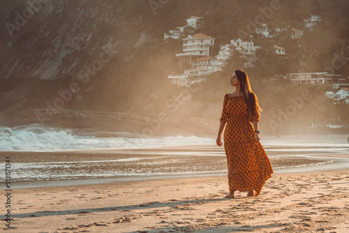 Beautiful brunette girl walking barefoot at the beach wearing a dress by sunset time photo