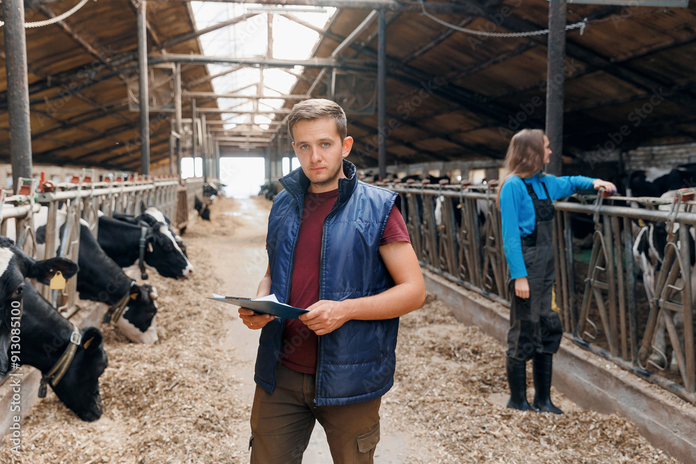 Portrait young man farmer with clipboard checking cows in cowshed on dairy farm, background vet doctor control health of cattle. Concept modern agriculture business industry, farming animal husbandry Portrait young man farmer with clipboard checking cows in cowshed on dairy farm, background vet doctor control health of cattle. Concept modern agriculture business industry, farming animal husbandry