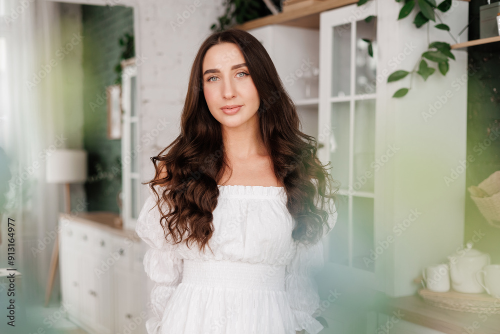 Women's day. Romantic attractive young woman 20-25 years with long curly hair in white dress is poses with crossed hands and looks at camera at modern stylish interior of home kitchen on background. Women's day. Romantic attractive young woman 20-25 years with long curly hair in white dress is poses with crossed hands and looks at camera at modern stylish interior of home kitchen on background.