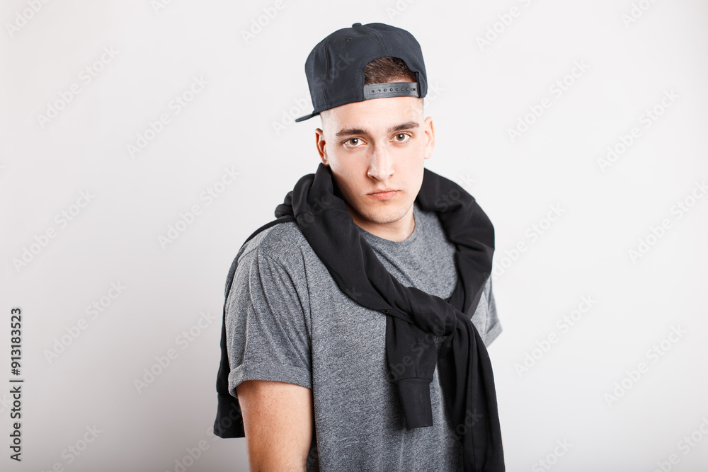 Handsome fashion man with cap in grey T-shirt with pullover on shoulders on white background in studio