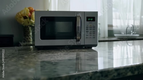 A silver microwave sits on a countertop in a kitchen. The microwave is turned off and has a digital display. The kitchen is well-lit and has a clean, modern look
