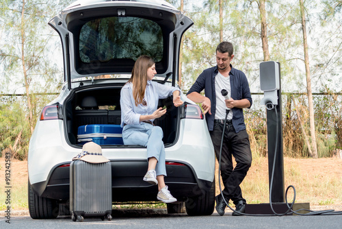 Lovely young couple recharging battery for electric car while relaxing and sitting on the trunk during road trip travel EV car in autumnal forest. Eco friendly travel on vacation during autumn. Exalt