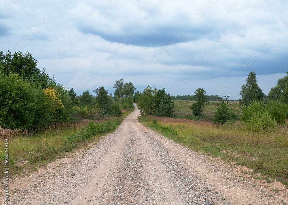 Gravel road in the countryside. Trees and meadows around, cloudy sky Gravel road in the countryside. Trees and meadows around, cloudy sky