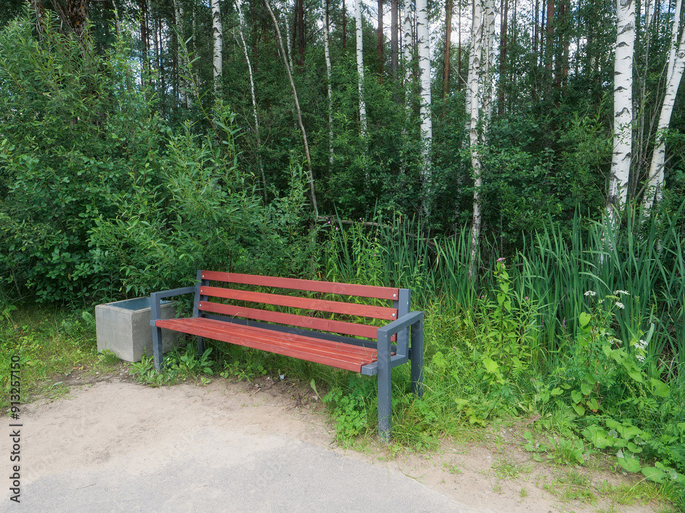 Bench in a park with metal frame and brown wood color plastic, rubbish bin on a side, trees and tall green grass in the background. Nobody. Bench in a park with metal frame and brown wood color plastic, rubbish bin on a side, trees and tall green grass in the background. Nobody.