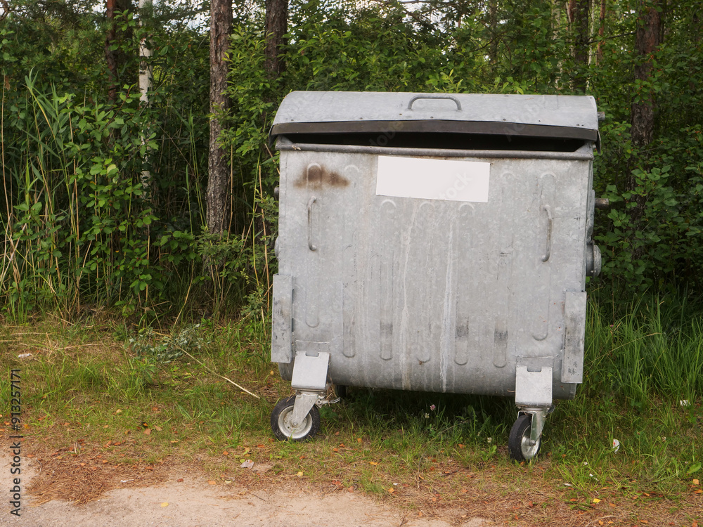 A large metal trash can sits on the side of a road. The trash can is closed and is surrounded by grass. The scene is peaceful and quiet, with no one around. Park maintenance and care. Waste business A large metal trash can sits on the side of a road. The trash can is closed and is surrounded by grass. The scene is peaceful and quiet, with no one around. Park maintenance and care. Waste business