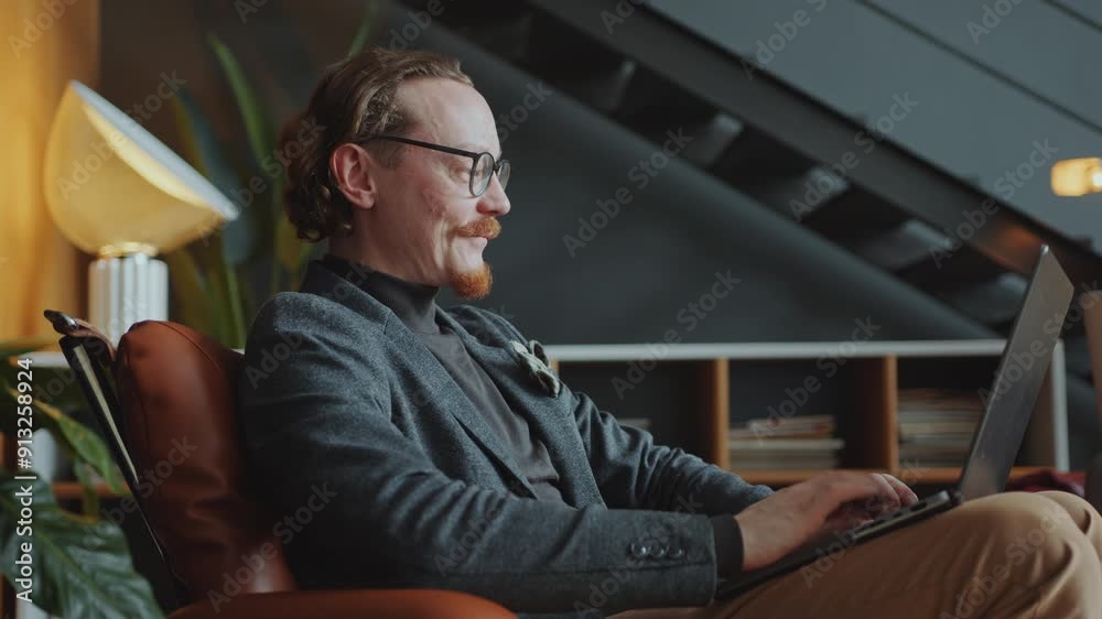 Caucasian businessman with beard and mustache wearing glasses and smart casual outfit sitting in armchair in the office and typing on laptop during workday. Side view Caucasian businessman with beard and mustache wearing glasses and smart casual outfit sitting in armchair in the office and typing on laptop during workday. Side view